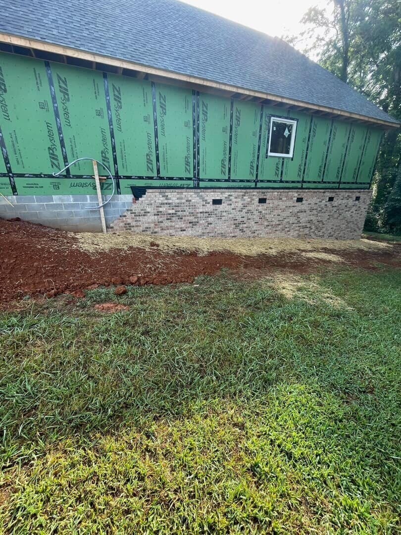 Exterior side view of a custom home under construction with green wrap and brick foundation by Debuty Builders, Knoxville, TN